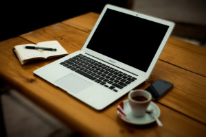 Macbook Air computer, with a writer's notebook, smartphone, and a cup of coffee, all on a wooden desk